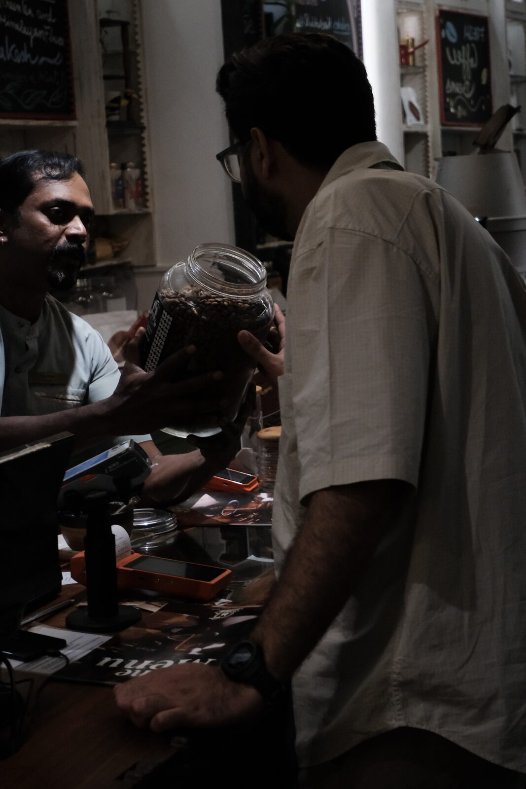 A barista handing a large glass jar of freshly roasted coffee beans across the counter, describing the beans with both hands.