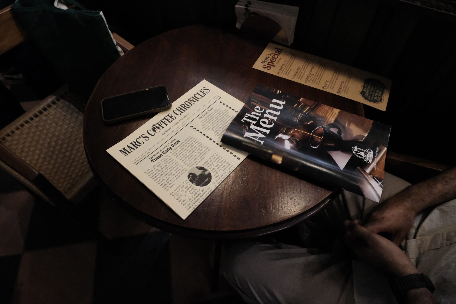 A wooden cafe table with the Marc's Coffee Chronicles newspaper, The Menu booklet, and Marc's Special specials card laid out.